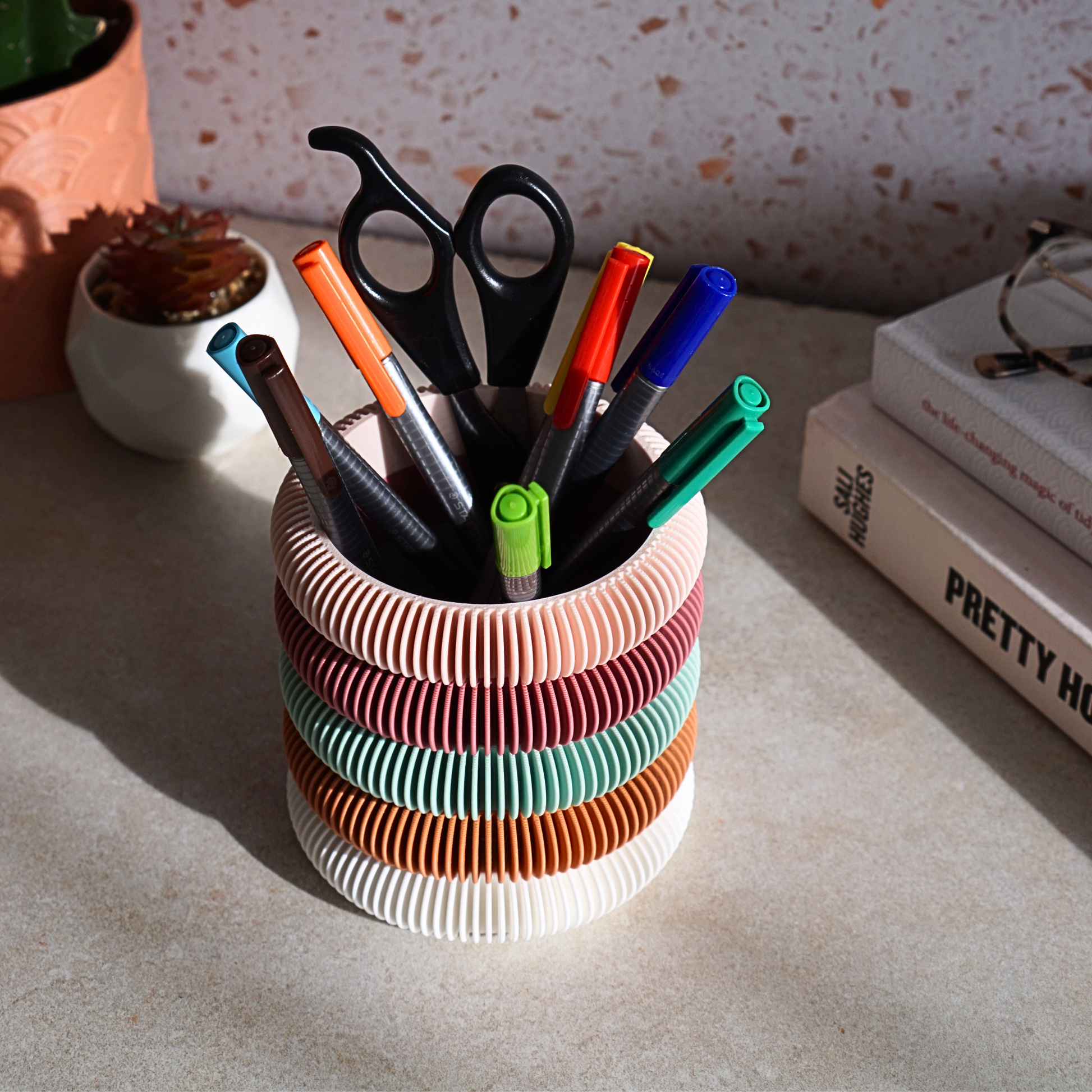 Bright coloured funky storage pot holding pens, pencils and scissors on a desk with plants and books. Home organisation and storage